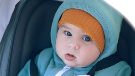Adorable funny boy in warm clothes sits in a stroller. Sweet grey-eyed kid looks at camera calmly. Close up.