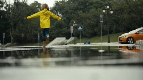 Close-up-a-happy-teenage-girl-in-a-yellow-jacket-and-rubber-boots-runs-through-a-puddle-splashing-water-to-the-sides-after-rain-in-the-park