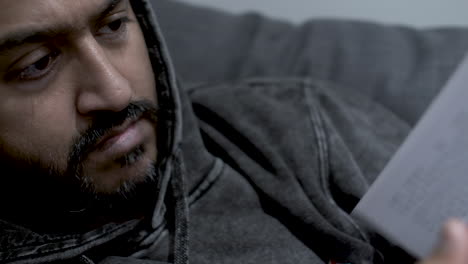 a close up shot of an indian man wearing a hoodie as he relaxes at the end of a long day, flicking through the pages of a book he is reading in search of a specific chapter