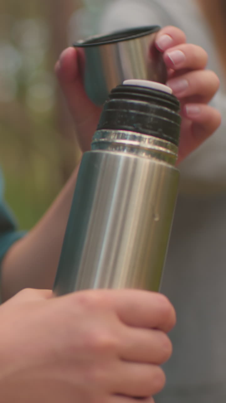primer plano de una mujer joven con camisa verde presionando la cubierta interior de un termo para acceder al agua en un bosque pacífico, mientras que otra mujer, ligeramente borrosa en el fondo, se cepilla el cabello