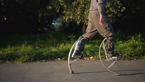 lower shot of man walking with stilts wearing gray pants on paved road surrounded by green grass and trees, showcasing focused motion, balance, and outdoor activity, emphasizing agility, creativity