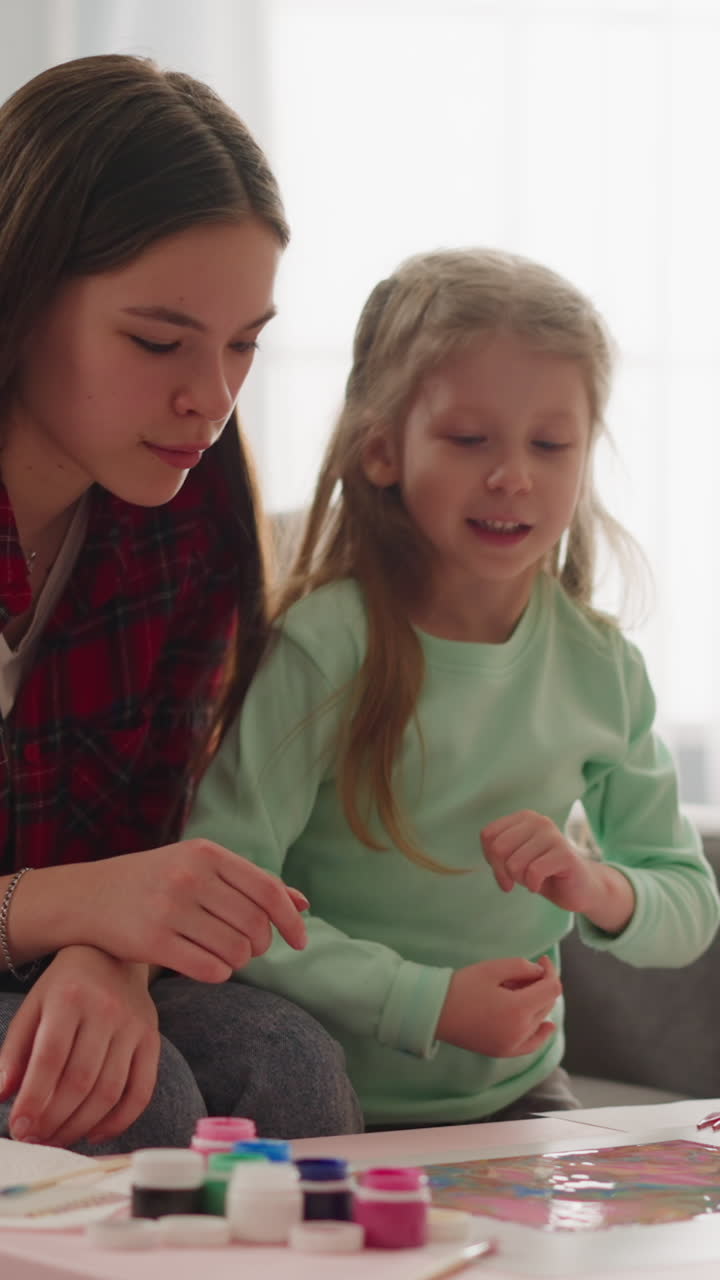 una niña rubia habla con una niñera feliz mirando un dibujo al estilo erbu sentada en una mesa en un apartamento en cámara lenta. lección de arte de mármol en casa