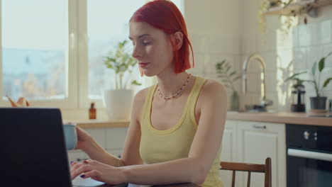 red-haired girl drinking coffee and working on laptop in cozy kitchen