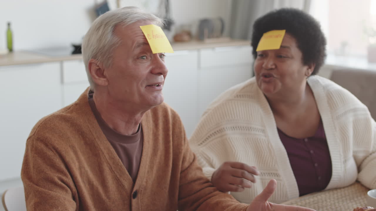 Tilting up of older people sitting at table, playing Forehead Detective game with yellow sticky notes on head, African woman talking and applauding Caucasian man winning, taking note off