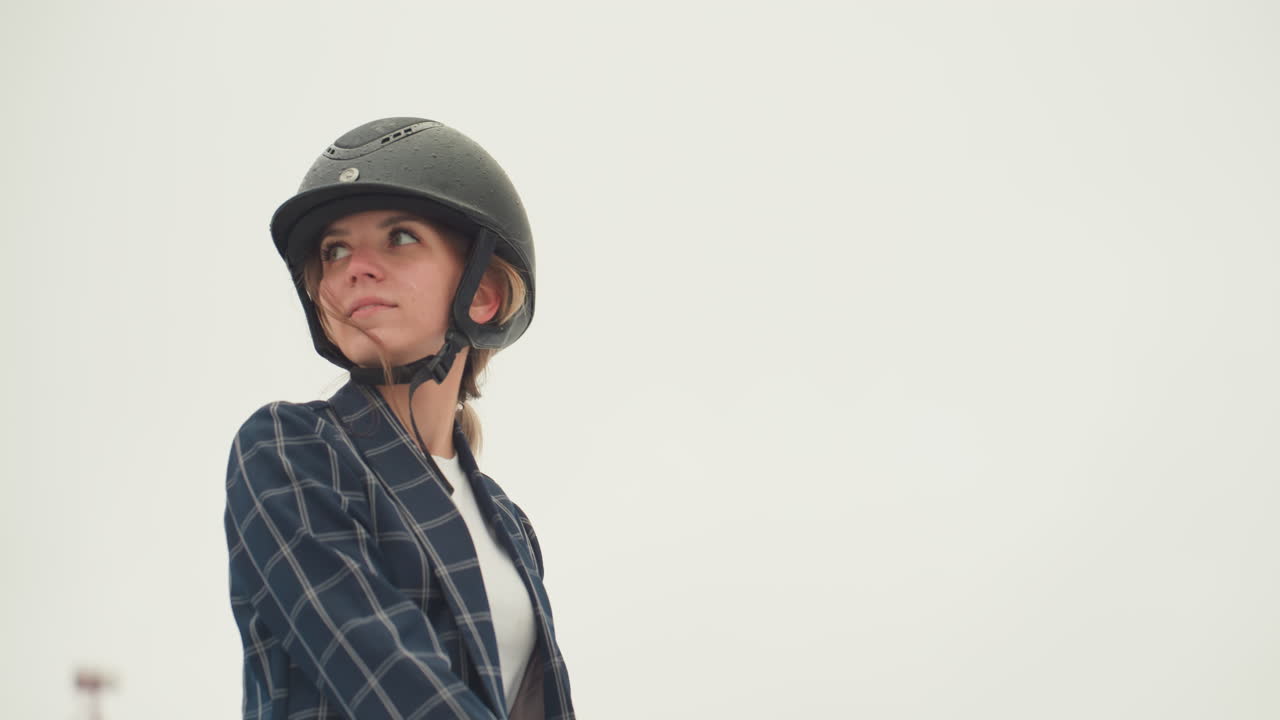 White woman in helmet looking sideways, focused posture and steady gaze, plaid blazer, equestrian education session, neutral sky background, closeup upper body, lesson atmosphere