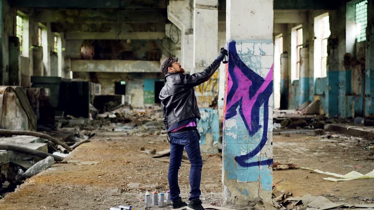 Graffiti painter in casual clothing is drawing with spray paint on column in spacious abandoned building. Abstract images, modern art, creative people and hipsters concept.