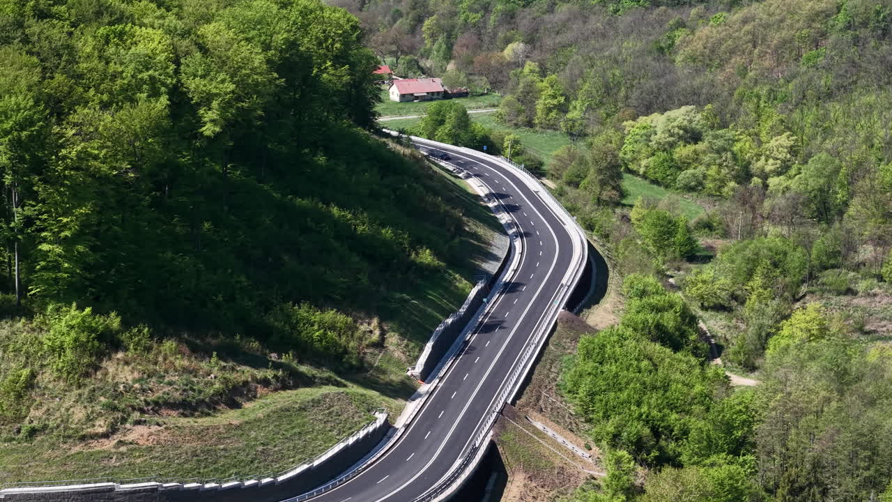 Una vista aerea cattura un'autostrada moderna e tortuosa che attraversa una lussureggiante foresta verde e una collina, simboleggiando lo sviluppo di nuove infrastrutture in un ambiente naturale.