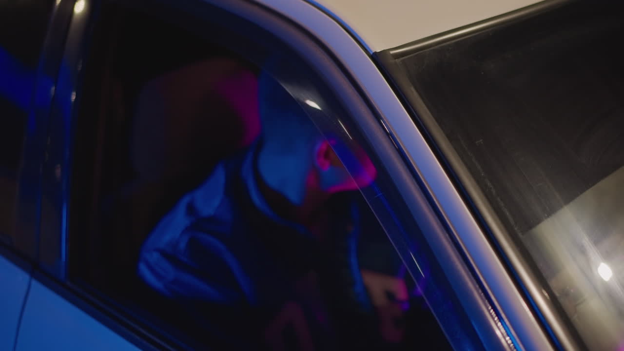 Giovane conducente in auto illuminata al neon guarda verso un dispositivo portatile all'interno di un veicolo parcheggiato, bagliore rosa-viola, luce interna del cruscotto, atmosfera tesa e introspettiva, tranquilla notte in città, fattorino che controlla