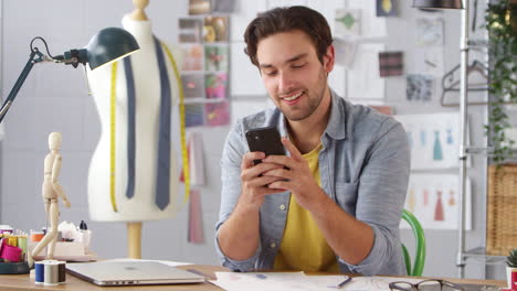 Male-Owner-Of-Fashion-Business-Using-Mobile-Phone-Sitting-At-Desk-In-Studio