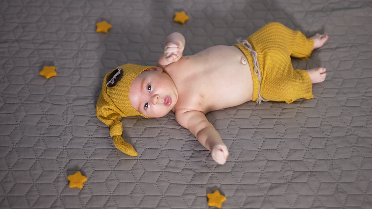 Infant child in a costume of gnome lies on the bed. Sweet boy waving his hands and turns away from the camera. View from above.
