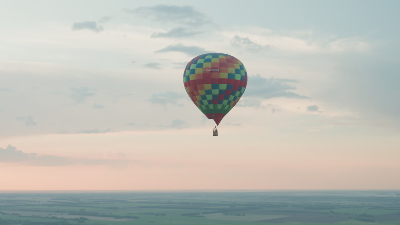 Colorful checkered hot air balloon carrying two passengers drifting above vast farmland and winding river at sunrise, distant serene flight against pastel sky
