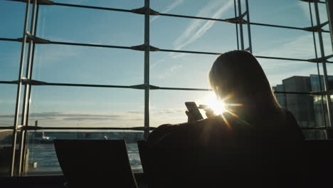 공항 터미널에서 큰 창을 마주하고 앉아 있는 여성의 실루엣은 스마트폰을 사용합니다.