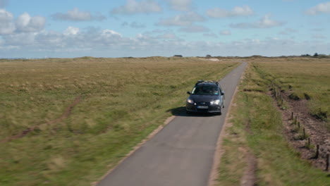 Slide-and-pan-shot-of-car-driving-on-narrow-asphalt-path-in-flat-landscape.-Backwards-tracking-of-vehicle-on-road-surrounded-by-vast-grasslands.-Denmark