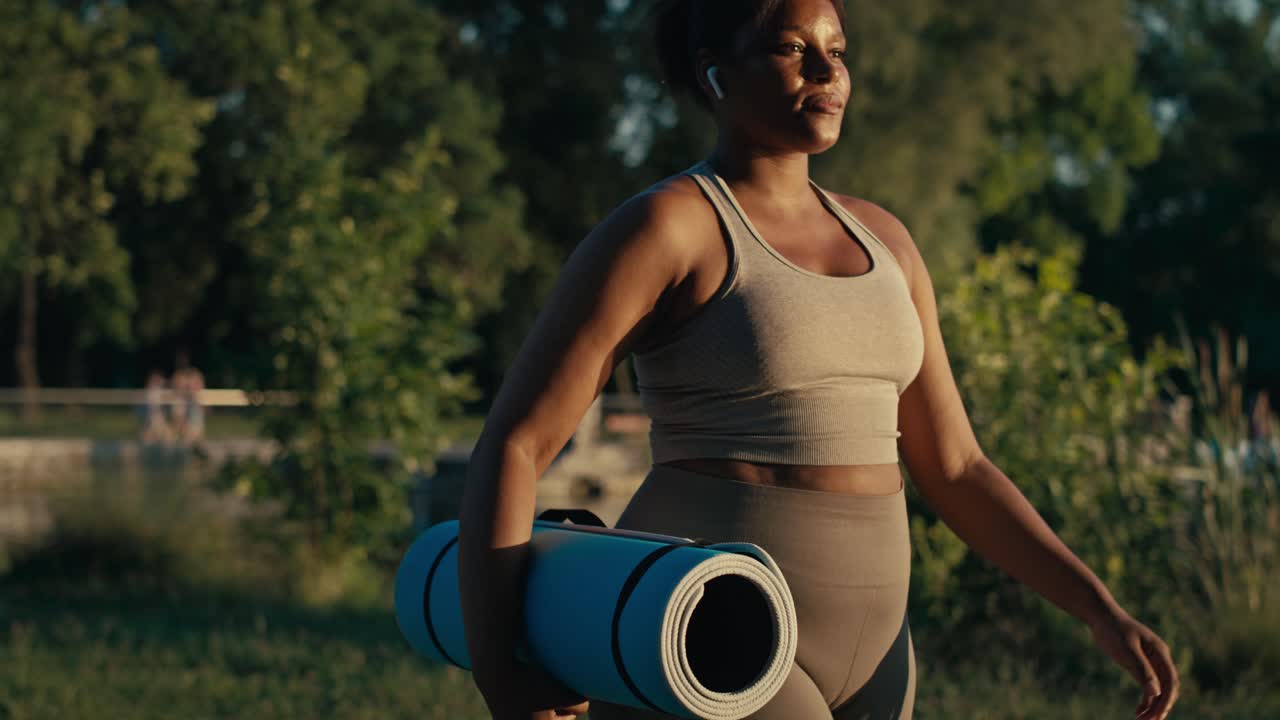 Plus sized African-American woman walking with exercise mat and earphones through the park in a summer day.