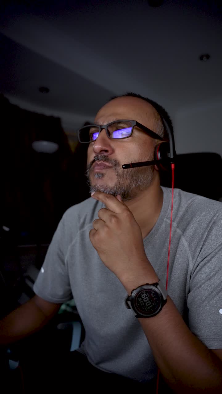 Bearded south asian man wearing headset and glasses talking and thinking during night shift at home