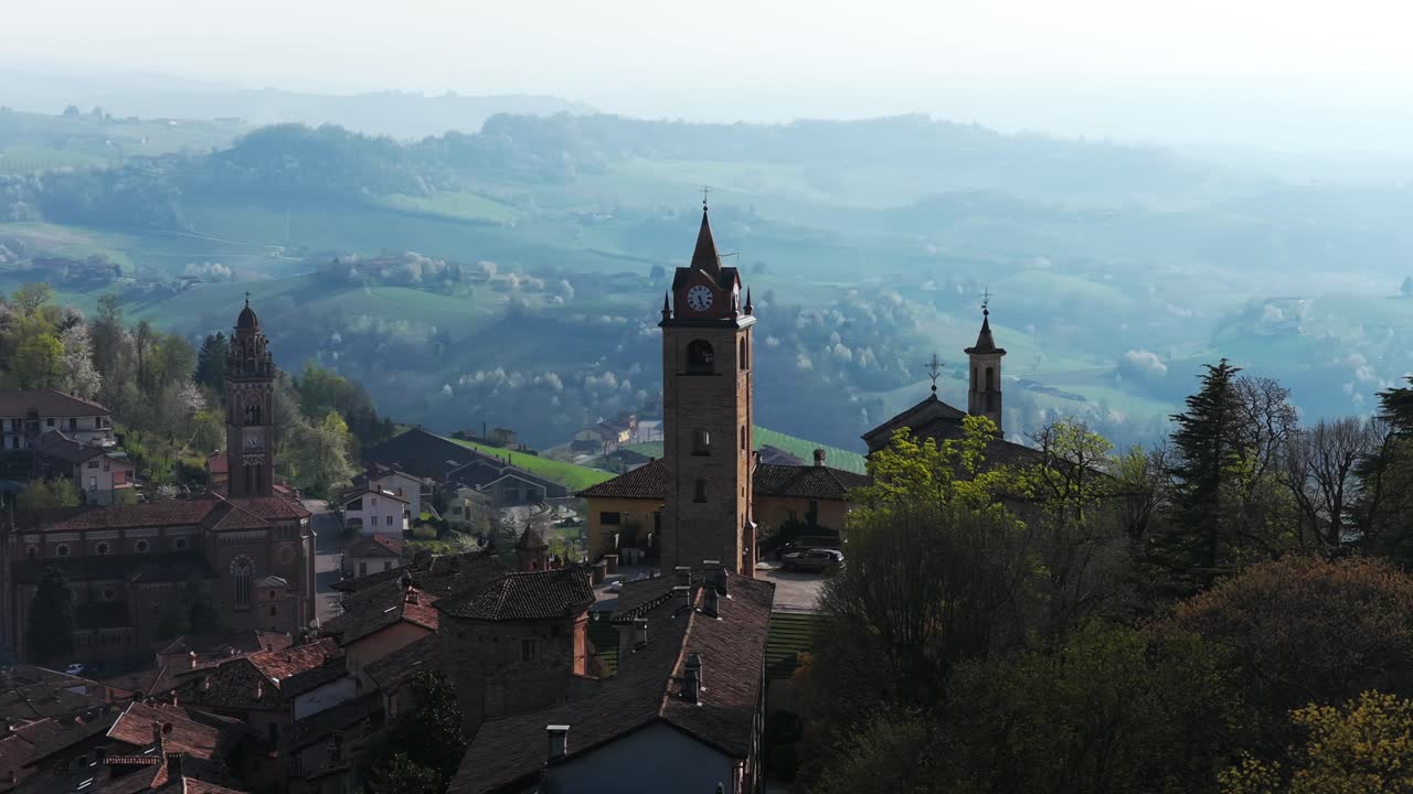 Monforte d'Alba at hazy sunrise, historic hilltop village with church towers, misty Langhe hills, Italian travel, wine region, Italy. Aerial drone orbiting