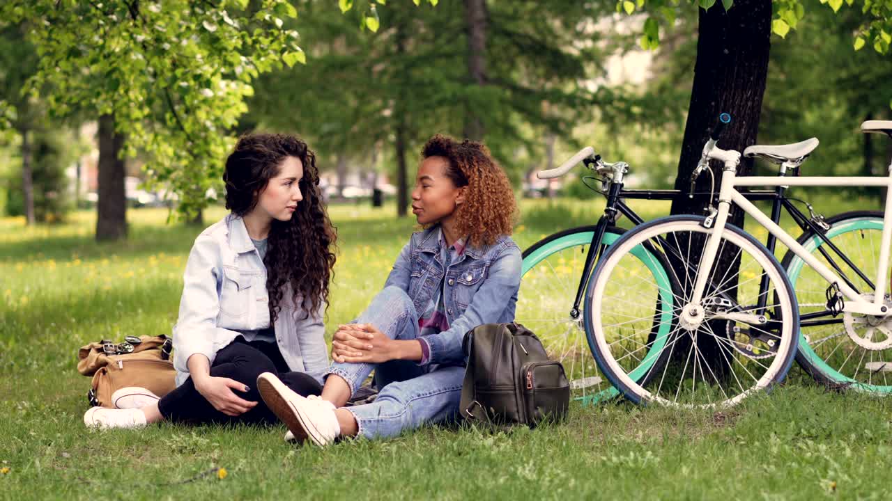una bonita mujer afroamericana está hablando con su amiga europea en el parque sentada en la hierba, las chicas están charlando y riendo. bicicletas y árboles están en el fondo, día soleado.