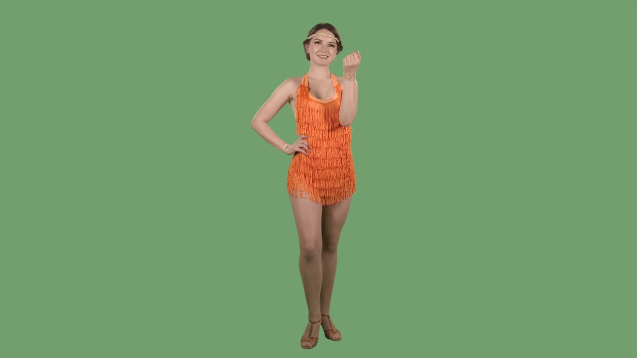 아름다운 여자가 카메라를 바라보고, 그녀의 손을 코하게 흔들고, 여기로 오십시오. 스튜디오에서 녹색 화면에 오렌지색 드레스와 프린지를 가진 갈색의 초상화.