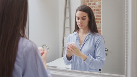 Frau-Trägt-Pyjamas-Zu-Hause-Im-Modernen-Badezimmer-Und-Verwendet-Feuchtigkeitscreme,-Die-Sich-Im-Spiegel-Spiegelt