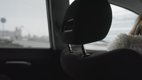side view of woman wearing white sweater seated inside car, waiting patiently for another vehicle to pass before driving, headrest partially visible, outdoor background