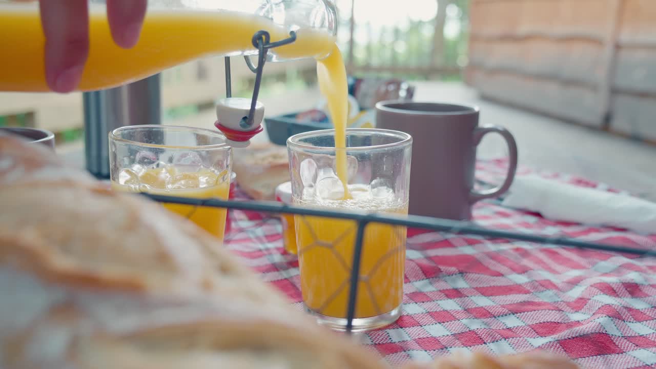 A person serving orange juice from a glass bottle into a drinking glass on a table set with baguette for a summer breakfast