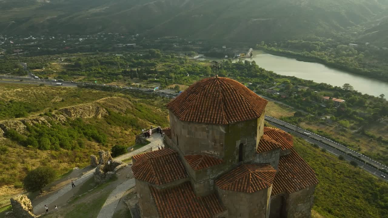 Jvari Manastırı'nın kırmızı kiremitli çatılarını ve taş yapısını gösteren, panoramik Gürcü manzarası ve tepeleriyle çevrili yakın bir drone görüntüsü