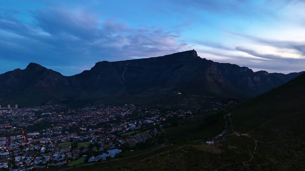 Veduta aerea cinematografica della Table Mountain a Città del Capo, Sudafrica, scogliere rocciose drammatiche con paesaggio urbano costiero al tramonto, nuvole che si muovono sulle cime e luce dorata della sera.