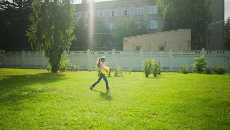 excited young girl walking briskly with yellow balloon under arm across bright grassy yard, sunlight creating warm glow on green field, with trees, bushes, and urban school building in background