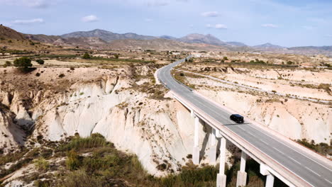 drone capturing a beautiful scenery of a black car driving over a bridge in the middle of a stunning landscape made out of mountains and a mixture of grassland and savanna, murica, spain