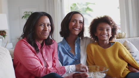 three generation female family group sit watching tv, laughing and eating popcorn, close up