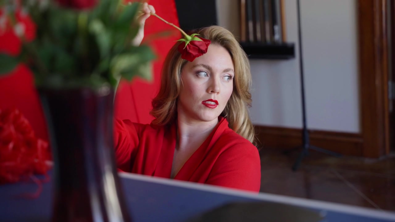 Kvinde i rød kjole arrangerer en rød rose indendørs, med bløde blomster i forgrunden, elegant makeup og en romantisk livsstilsatmosfære