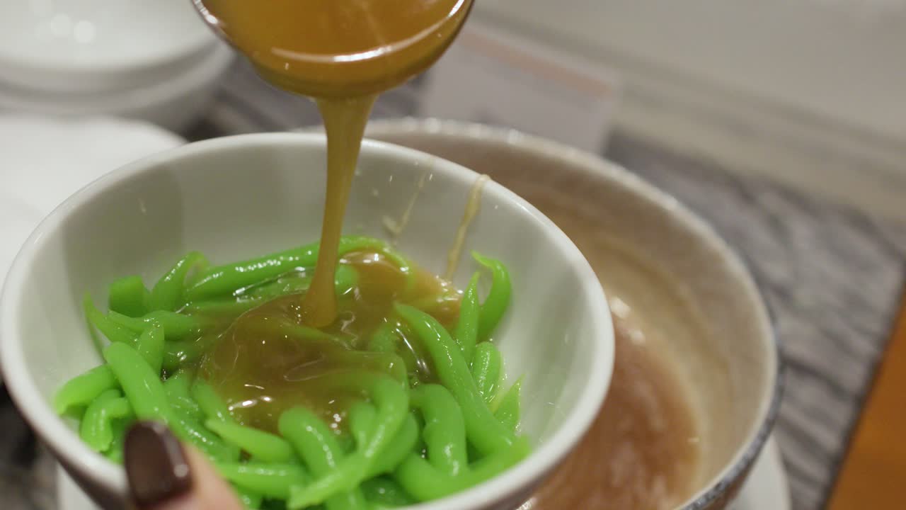 Close-up shot of brown palm sugar syrup poured onto green pandan noodles in white bowl