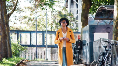 fashionable young black woman wearing a hat, sunglasses, an unbuttoned yellow pea coat and jeans walking along a treelined street looking around and using smartphone, low angle