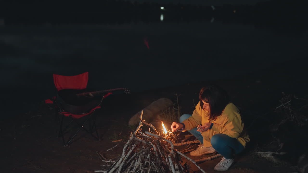 Nattecamping på stranden, solo campist tænder bål nær rød stol, arrangerer brænde og udstyr ved vandkanten, blide bølger i det fjerne, lanterne klar, reflekterende udendørs stemning og omhyggelig klargøring af lejrpladsen