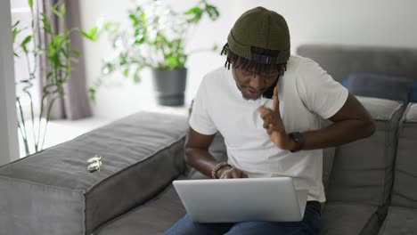 Afrikanischer-Mann-Benutzt-Smartphone,-Während-Er-Laptop-Auf-Dem-Sofa-Benutzt