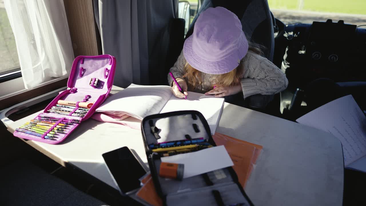 una niña con un sombrero rosa trabaja en su tarea escolar en una mesa dentro de una caravana iluminada por el sol junto a la ventana