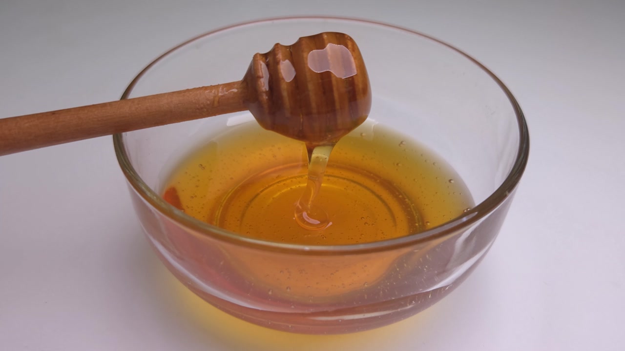 A clear glass bowl filled with honey. A wooden honey dipper tool is used to move and swirl the honey around, pick up the viscous liquid and lets it drip down.