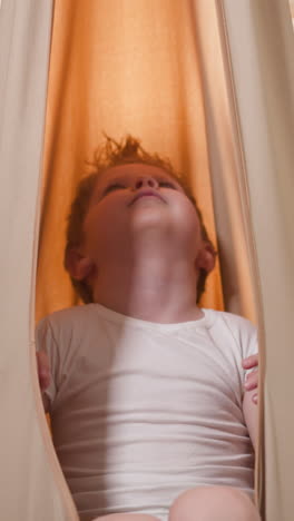 curious blond boy swaying in comfortable beige hammock looking at ceiling. cute little child in t-shirt enjoys relaxing in modern home gym slow motion