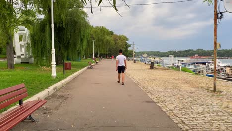 A-man-strolls-along-a-cliff-edge,-and-several-boats-are-docked-on-the-peaceful-riverside