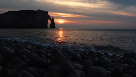 Wide-show-showing-splashing-ocean-water-on-stony-beach-and-silhouette-of-rocky-cliffs-during-sunset-in-background