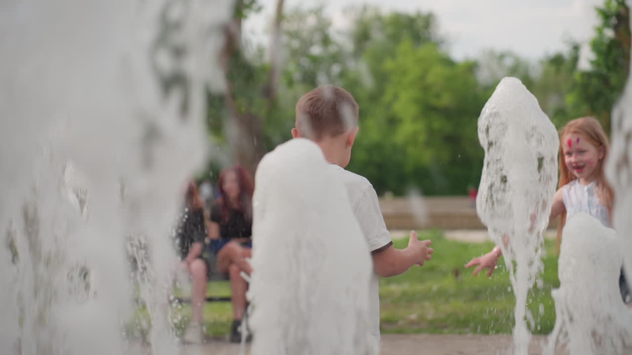 Excited toddlers playing with fountain water in recreational park, laughing and splashing near bench with seated adults blurred in background, summer greenery framing joyful playful interaction