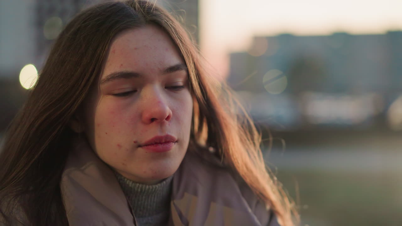 un primer plano de una mujer joven con una chaqueta de melocotón, que parece perdida en profundos pensamientos con una débil sonrisa en su rostro. el fondo urbano borroso y la suave luz de la tarde