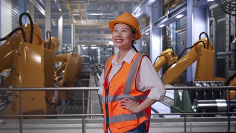 side view of asian female engineer wearing safety helmet looking around while standing with arms akimbo with industrial robotic arms working