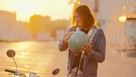 Porträt-Eines-Glücklichen-Kerls-Mit-Langen-Lockigen-Haaren-In-Einem-Jeanshemd,-Der-Seinen-Grünen-Mopedhelm-Vom-Kopf-Nimmt,-Lächelt-Und-An-Einem-Sonnigen,-Hellen-Morgen-Bei-Sonnenaufgang-Neben-Seinem-Moped-In-Die-Kamera-Schaut