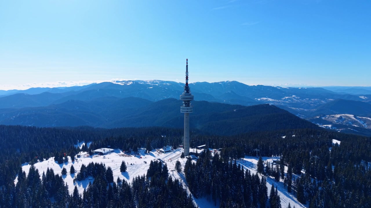 Torre de telecomunicações no topo de uma montanha nevada acima de florestas de pinheiros. Uma estrutura alta de concreto ergue-se entre árvores verde-escuras sob um céu azul