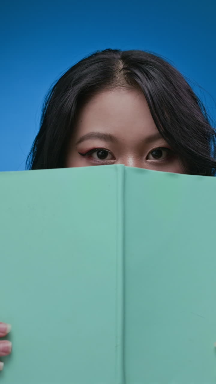 Asian Student with Book Posing for Camera on Neon Blue Wall Background