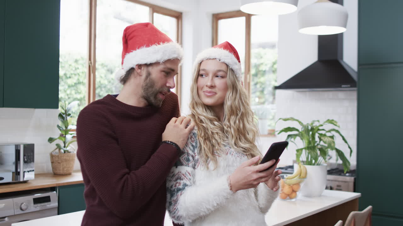 una pareja diversa con sombreros de santa usando un teléfono inteligente en la cocina, en cámara lenta