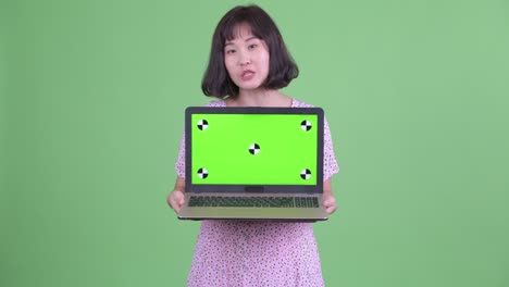 Studio shot of beautiful pregnant Asian woman showing copy space from laptop against green studio background