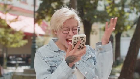 happy caucasian blonde woman using smartphone celebrating win good message news on city town street