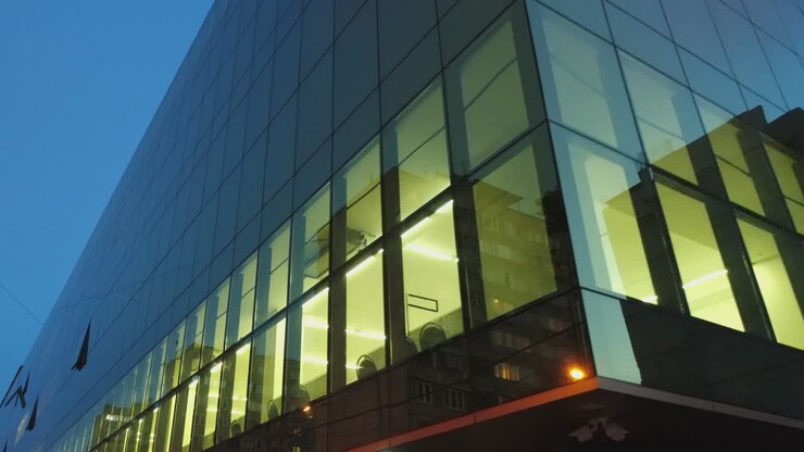 Vista esterna 4K di un edificio nel quartiere degli affari, centro commerciale con finestre a specchio di sera o di notte. Riflesso di città, persone, auto nelle pareti a specchio. Telecamera drone in movimento. Persone che lavorano all'interno.
